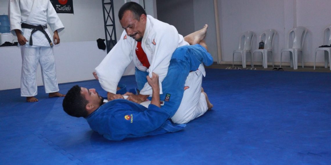 Atleta vestindo um kimono branco, aplica um golpe enquanto está por cima de seu oponente, que veste um kimono azul e está de costas sob o tatame - Foto: Divulgação